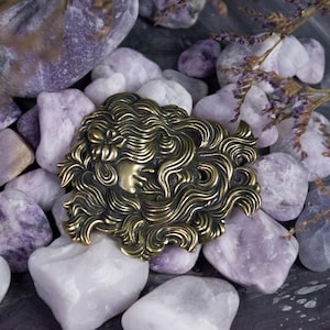 May include: A gold-toned metal brooch with a detailed design of a woman's face with flowing hair. The brooch is surrounded by purple and white stones.