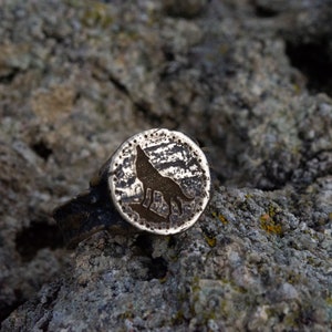 May include: A silver ring with a wolf silhouette design. The ring is textured and has a circular shape.
