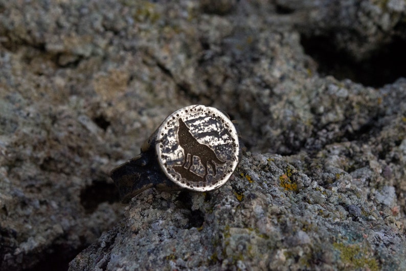 May include: A silver ring with a wolf silhouette design. The ring is textured and has a circular shape.