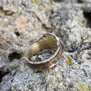 May include: A gold-colored ring with a worn, textured surface. The ring is lying on a rough, gray rock.