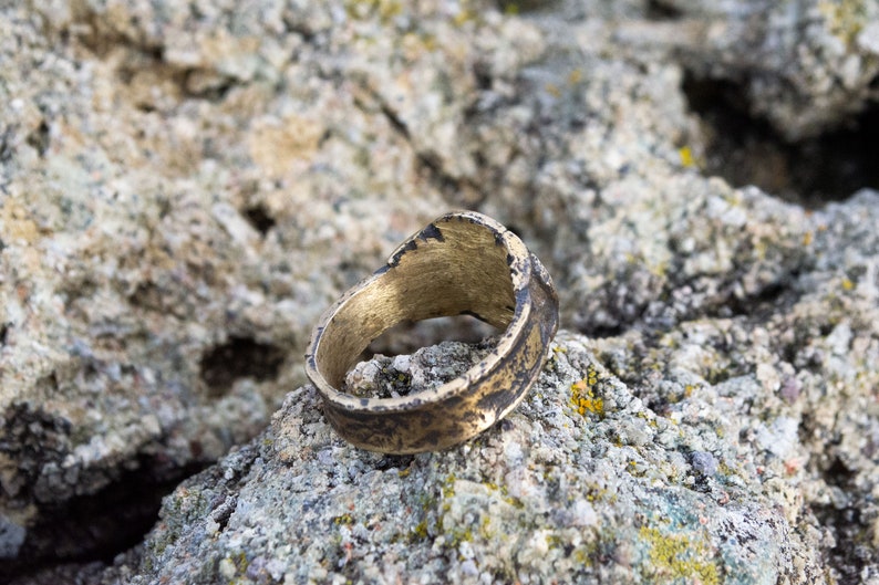 May include: A gold-colored ring with a worn, textured surface. The ring is lying on a rough, gray rock.