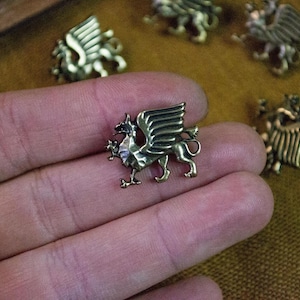 Può includere: Una piccola spilla metallica color bronzo a forma di grifone con ali spiegate e coda rivolta verso l'alto. La creatura è raffigurata di profilo, mostrando piume alari dettagliate e un becco appuntito. La spilla ha una finitura liscia e riflettente.