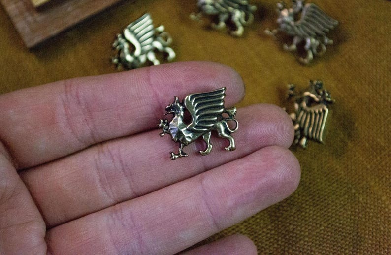 May include: A small, metallic, bronze-colored pin shaped like a griffin with wings extended and a tail curled upward. The creature is depicted in profile, showing detailed wing feathers and a sharp beak. The pin has a smooth, reflective finish.