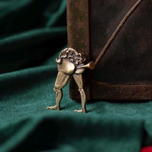 May include: A quirky brass pendant depicting a figure playing a trumpet. The figure has a large head and legs, with the trumpet emerging from its rear. The pendant is set against a dark green and red backdrop.