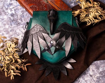 May include: A teal leather bag with a raven design. The bag has black wings and a raven head with a beak. The bag is on a brown fabric background.