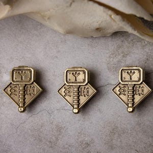 Peut inclure: Trois pendentifs de couleur bronze avec un motif de marteau. Chaque pendentif a un dessus rectangulaire avec un symbole, une base en forme de losange et une poignée verticale. Ils sont disposés sur une surface grise texturée.