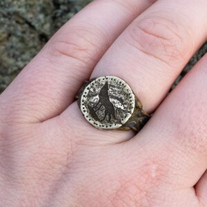 May include: A silver ring with a circular design featuring a wolf silhouette. The ring is worn on a finger.