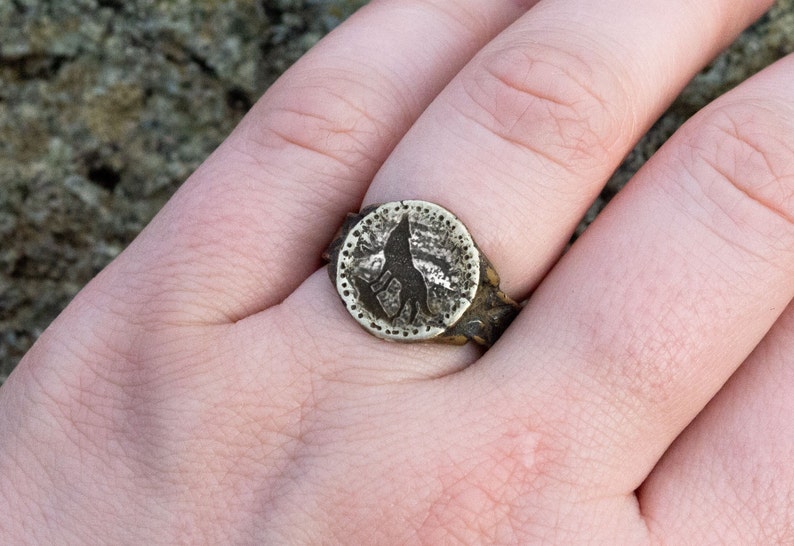 May include: A silver ring with a circular design featuring a wolf silhouette. The ring is worn on a finger.