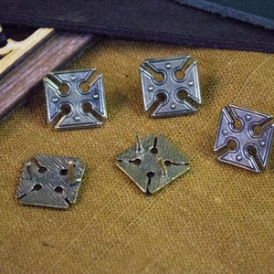 May include: Five silver-toned metal buttons with a cross design. The buttons have a raised, textured surface and are arranged on a brown fabric background.