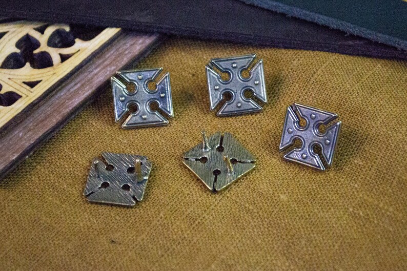 May include: Five silver-toned metal buttons with a cross design. The buttons have a raised, textured surface and are arranged on a brown fabric background.