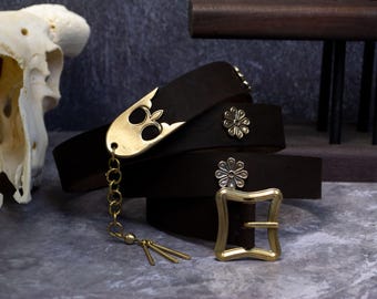 May include: Dark brown leather belt with a gold-colored buckle and decorative accents. The belt features a unique gold-colored pendant with a chain and tassels. The buckle is square with rounded edges. The belt is approximately 1.5 inches wide.