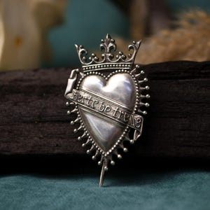 May include: A silver-toned metal brooch shaped like a heart topped with a crown. A banner wraps around the center of the heart, featuring the engraved text 'herte be trewe'. The heart is surrounded by a radiating sunburst of small metal spikes.