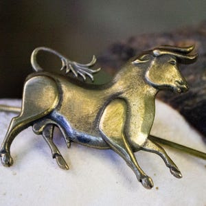 May include: A gold-toned metal bull figurine brooch with a detailed design. The bull is running with its head turned to the side. The brooch is attached to a long, thin metal pin.