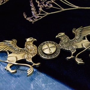 Pode incluir: Um spinner de metal dourado com duas figuras de grifos com as asas abertas. O spinner tem um disco central rotativo com um design de cruz.