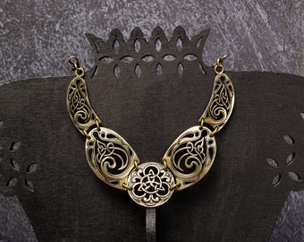 Puede incluir: Un collar dorado con un intrincado diseño de nudos celtas. El collar se exhibe sobre un soporte negro con un diseño decorativo recortado. El collar presenta un colgante central circular y piezas laterales curvas.