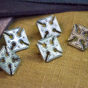 May include: Five silver and bronze metal decorative buttons with a cross design. The buttons are on a brown fabric surface.