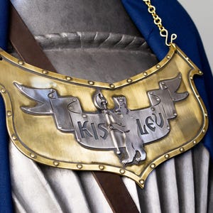 May include: A brass and silver shield-shaped pendant with the text "KISLEV" and a bear illustration. The pendant is attached to a gold chain and is set against a blue fabric background.