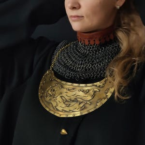 May include: A gold-colored metal necklace with a decorative design. The necklace is worn over a black chainmail collar.