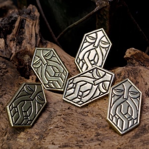 Peut inclure: Cinq épingles géométriques métalliques avec un motif de visage stylisé. Les épingles sont dans des tons argent et bronze, dispersées sur une surface en bois brun texturé. Les épingles ont un aspect artistique unique.