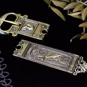 May include: A brass belt buckle and matching strap end featuring intricate relief carvings. The buckle displays a skull motif, while the rectangular strap end depicts a full-length skeletal figure. Both pieces have an aged, metallic finish.
