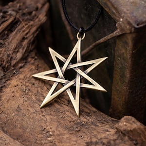 Puede incluir: Un collar con colgante de color bronce que presenta un diseño de estrella de siete puntas. La estrella está suspendida de un cordón negro y descansa sobre una superficie de madera marrón texturizada. El colgante tiene un diseño geométrico calado.