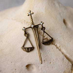 May include: A bronze-toned metal brooch shaped like a sword with a crossguard that supports two hanging scales. The sword hilt and the points where the scales attach to the crossguard are accented with small, clear, sparkling inset stones.