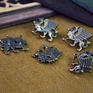 May include: A set of five silver-toned metal pins shaped like winged griffins. Each pin features a detailed relief of the mythical creature with prominent wings and legs. Two of the pins are positioned to show their flat backs with two metal posts.