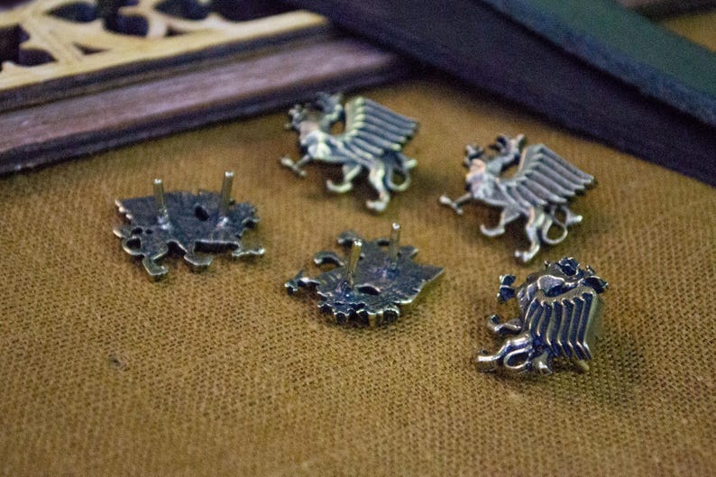 May include: A set of five silver-toned metal pins shaped like winged griffins. Each pin features a detailed relief of the mythical creature with prominent wings and legs. Two of the pins are positioned to show their flat backs with two metal posts.