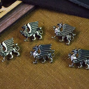 May include: Five silver-toned metal buttons, each shaped like a griffin with wings spread, a long tail, and a detailed body. These metallic fasteners feature a polished finish and a raised, three-dimensional design depicting the mythical creature.