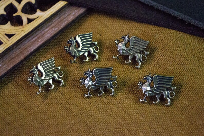 May include: Five silver-toned metal buttons, each shaped like a griffin with wings spread, a long tail, and a detailed body. These metallic fasteners feature a polished finish and a raised, three-dimensional design depicting the mythical creature.