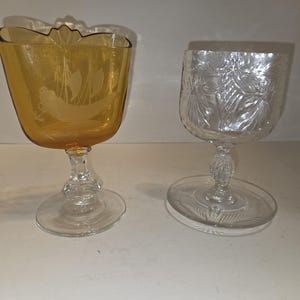 May include: Two vintage stemmed glasses. One is amber-colored with a sailboat etching, the other is clear with a floral pattern. Both have a decorative stem and a round base. Ideal for serving beverages.