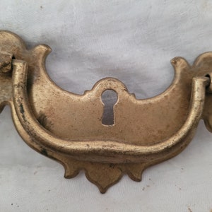 May include: Antique brass drawer pulls with a keyhole. The pulls have a curved design with decorative flourishes. The brass has a weathered appearance, suggesting age. These pulls are suitable for vintage furniture restoration projects.