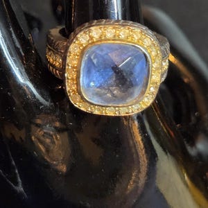 May include: A ring featuring a square-cut blue gemstone set in a gold-toned bezel, surrounded by a halo of small clear stones. The band is silver-toned with a textured, ridged pattern along the sides, providing a contrast to the central setting.