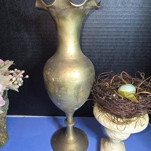 May include: A vintage brass vase with a ruffled rim. The vase has a bulbous body and a flared base. The vase is approximately 10 inches tall. A decorative bird's nest with an egg sits nearby.