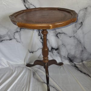 May include: A brown wooden pedestal table featuring an octagonal top with a decorative embossed border. The table stands on a central turned column supported by three curved legs, showcasing a polished finish and visible wood grain details.