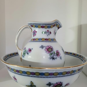 May include: White porcelain pitcher and basin set. The pitcher has a curved handle and spout, and sits inside the basin. Both pieces are decorated with pink and green floral patterns, butterflies, and gold trim.