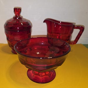 May include: A ruby red glass serving set, including a covered sugar bowl, a creamer, and a pedestal bowl. The pieces have a textured, geometric design. The set is displayed on a yellow surface.