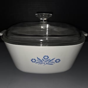 May include: White casserole dish with a clear glass lid and a clear knob. The dish has a blue floral design on the front. The dish has two small handles on either side.