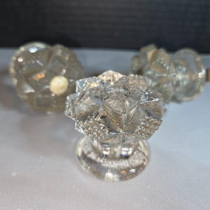 May include: Three clear glass bottle stoppers with faceted, starburst-like designs. The stoppers have a round base and a decorative top. The stoppers are made of clear glass and are likely vintage or antique.