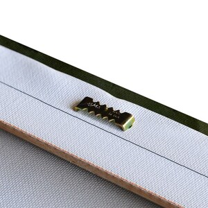 May include: Close-up of a gold-colored metal hanging hook attached to the back of a canvas painting.