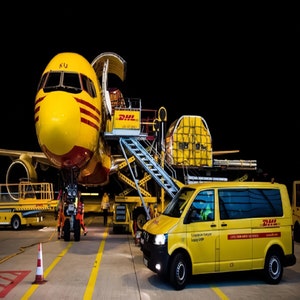 May include: A yellow DHL cargo plane with its cargo door open is being loaded by a yellow DHL van. The plane is parked on a tarmac with a yellow loading ramp and a yellow DHL ground crew vehicle.
