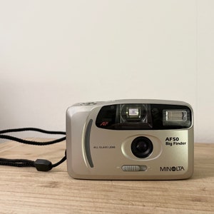 May include: A silver Minolta AF50 Big Finder 35mm film camera with a black strap. The camera has a built-in flash and an all-glass lens.