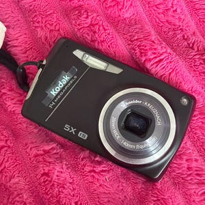 May include: A black Kodak digital camera with 14 megapixels and a 5X IS lens. The lens is labelled Schneider-Kreuznach 28mm WIDE-140mm (Equivalent). The camera features a silver flash unit and a black wrist strap attached to the side of the body.