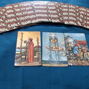 May include: A tarot card spread on a blue surface. The deck's backs have a red and white floral design. Three cards are face up, showing illustrations. The Ace of Cups card is visible, along with other cards from the deck.
