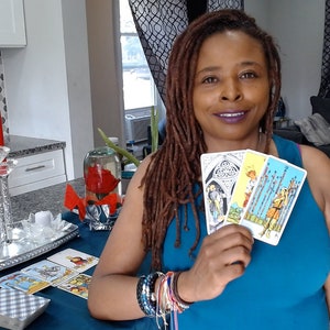 May include: A woman holding three tarot cards with colourful illustrations. A table displays a deck of cards and a decorative centrepiece with red candles. The woman wears a blue top and multiple bracelets.