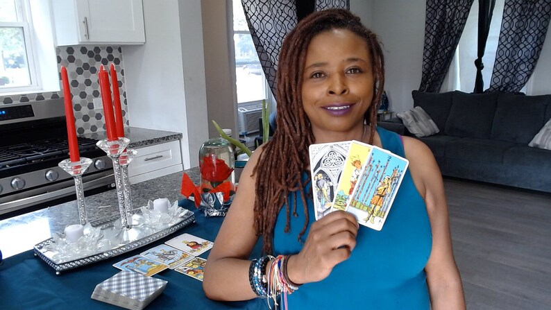 May include: A woman holding three tarot cards with colourful illustrations. A table displays a deck of cards and a decorative centrepiece with red candles. The woman wears a blue top and multiple bracelets.