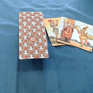 May include: A tarot card deck with a patterned back showing a repeating design of red and white floral motifs and a central rose. Three cards are fanned out, depicting figures with coins and staffs, on a blue cloth surface.