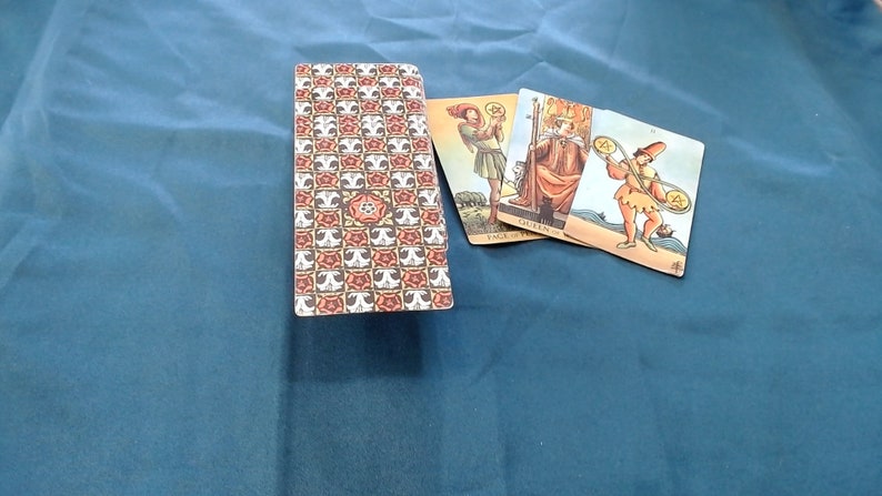 May include: A tarot card deck with a patterned back showing a repeating design of red and white floral motifs and a central rose. Three cards are fanned out, depicting figures with coins and staffs, on a blue cloth surface.