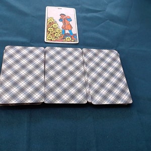May include: A tarot card spread with a card depicting a figure and gold coins, and three face-down cards with a blue and white plaid design. The cards are arranged on a dark teal surface.