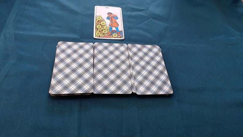 May include: A tarot card spread with a card depicting a figure and gold coins, and three face-down cards with a blue and white plaid design. The cards are arranged on a dark teal surface.
